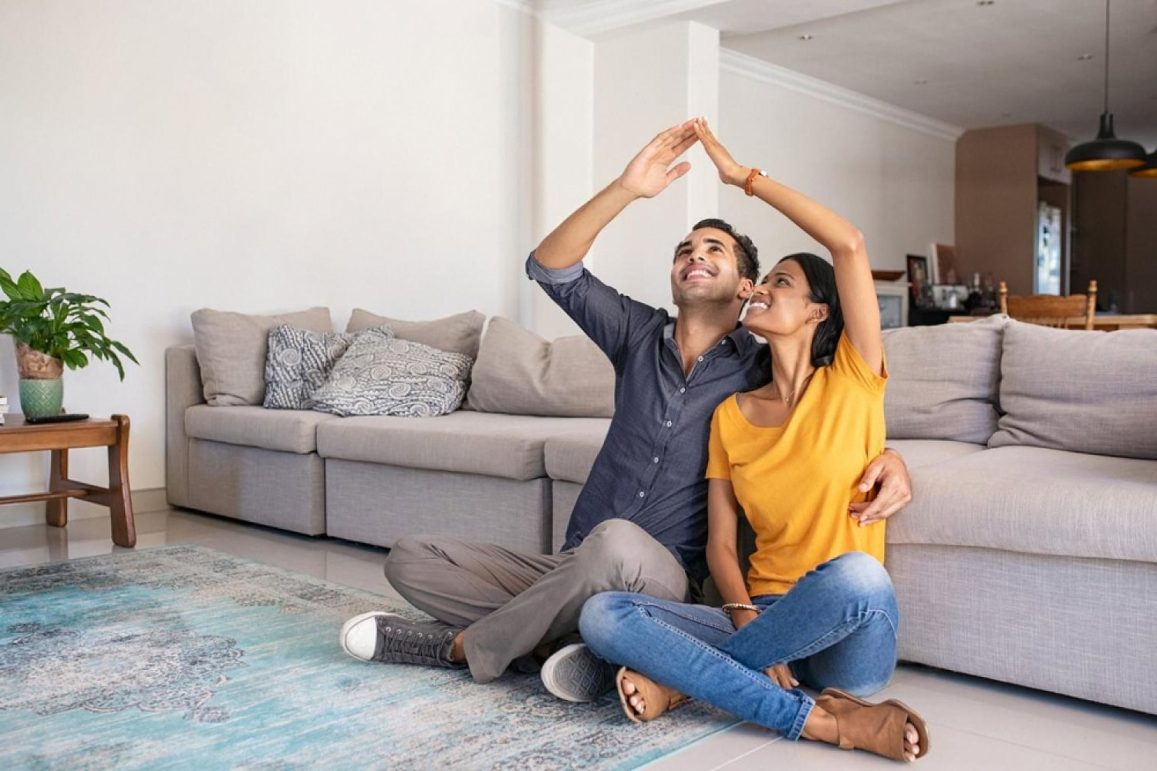 Young couple sitting on the floor of their UAE living room forming a roof shape with their hands, symbolising home insurance protection