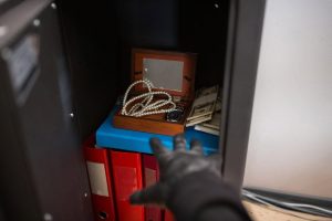Gloved hand reaching into a luxury home safe holding cash, a watch and a pearl necklace