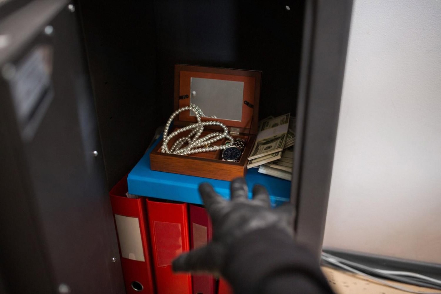 Gloved hand reaching into a luxury home safe holding cash, a watch and a pearl necklace